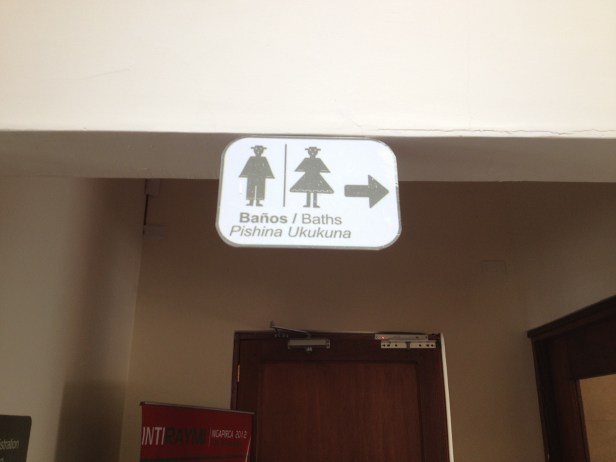 Placa em espanhol, inglê e quechua indicando banheiros. Olha os ícones representando homens e mulheres em trajes típicos, que bonitinho!